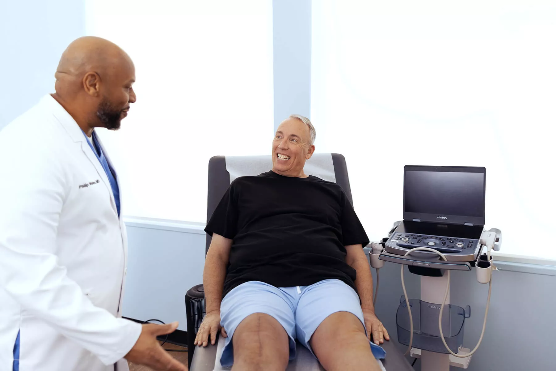 Vein specialist talking with a smiling patient seated in an exam chair next to an ultrasound machine