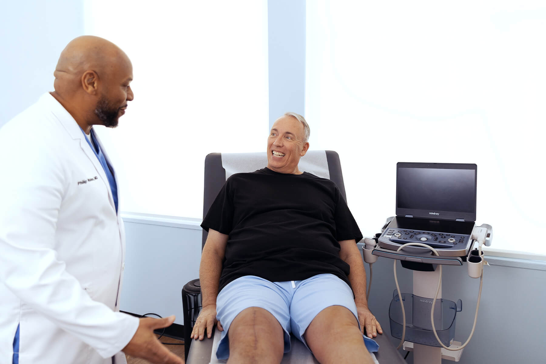 Vein specialist talking with a smiling patient seated in an exam chair next to an ultrasound machine